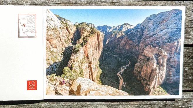 A postcard on a rustic wooden surface with an image of a scenic canyon. On the left side of the postcard, there is a small map marking the location of LOGE with the text "LOGE" and "Est. 2017."