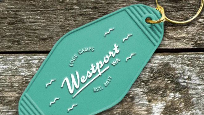 A close-up image of a teal keychain with a gold key ring, featuring the LOGE Camps logo and the text "Westport WA EST. 2017" embossed in white, and placed on a rustic wooden surface.
