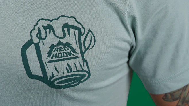 A close-up image of a person wearing a light gray T-shirt with a graphic of a frothy beer in a wooden mug featuring the Red Hook logo.